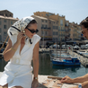 Woman in a white dress and headscarf by a marina with boats and buildings in the background.